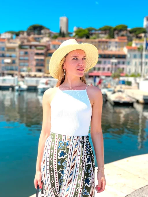 A girl with a hat, white to p and green skirt is looking to the right. Behind is a row of buildings. Picture is taken in Cannes as one of the best cities to stay in the French Riviera.